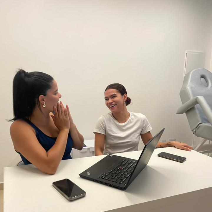 ´Duas médicas em primeiro plano conversando com os braços apoiados em uma mesa e ao fundo uma parede